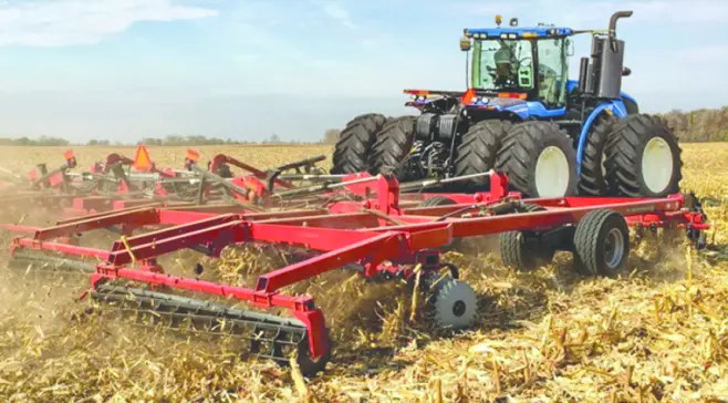 Blue row-crop tractor with large flotation ag tires pulling a red field cultivator through a harvested cornfield under a clear sky, illustrating agricultural tire and wheel use for farm equipment.