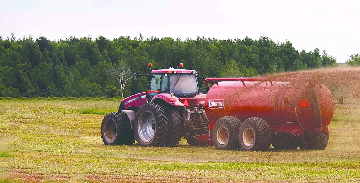 seasonal pic Red tractor with oversized agricultural tires pulling a red slurry tank spreading manure across a grassy Central Wisconsin field with tree line in background, illustrating farm tires, ag wheels, and on‑farm service for agricultural equipment.