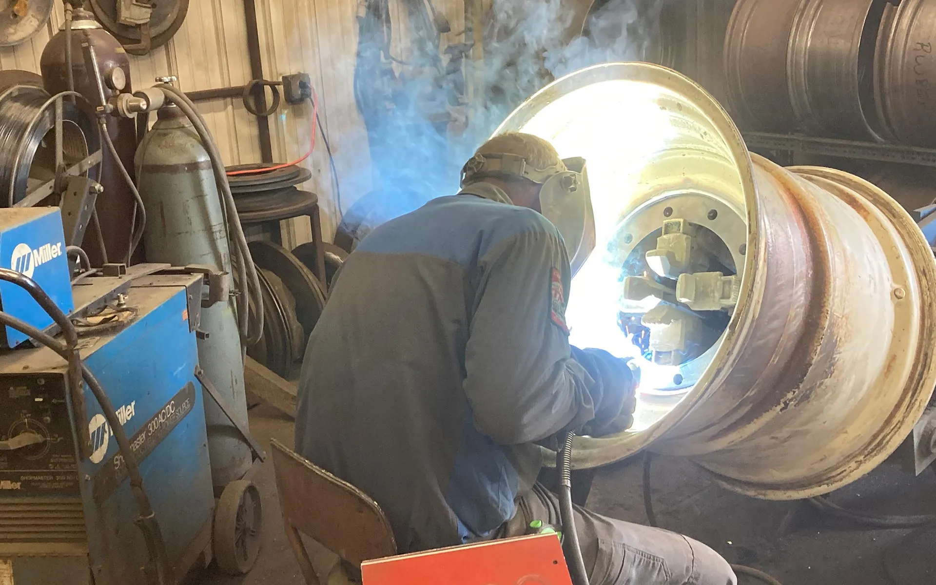 A shop technician in a welding mask welding the inside of a large rusted agricultural wheel rim indoors, surrounded by welding equipment, gas tanks, and stacks of tires and wheels at Bill’s Tire & Service in Colby, WI