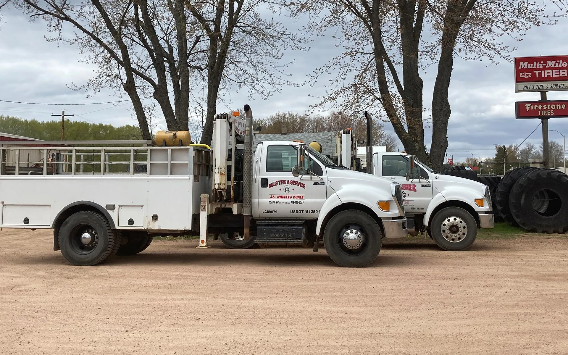 Two white on‑farm service trucks parked on a gravel lot in front of large farm tires and a Multi‑Mile/Firestone sign, with Bill’s Tire & Service logos visible on the doors and leafless trees and cloudy sky in the background
