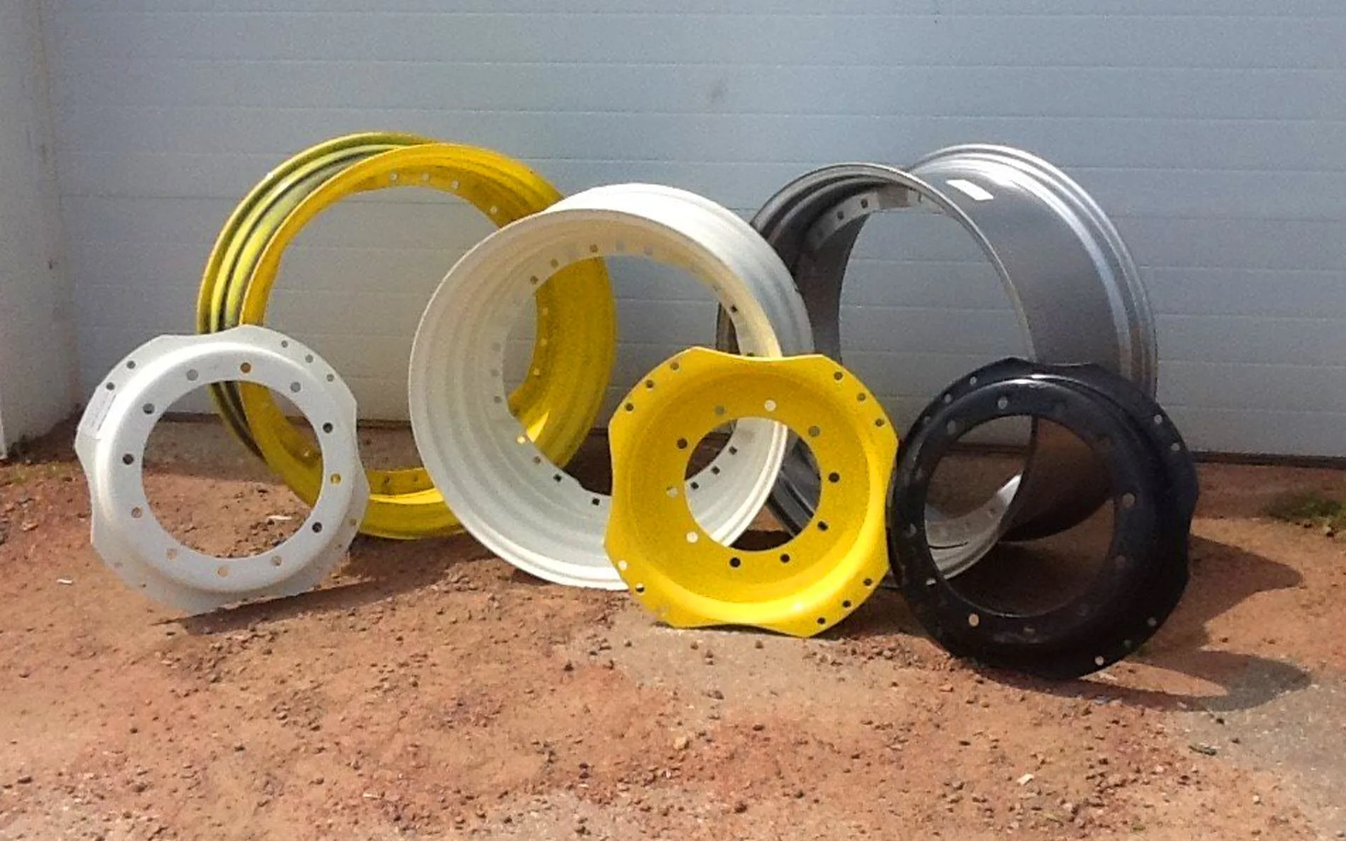 Several large painted tractor and implement wheel rims in white, yellow, gray and black leaning against a garage door on a dirt lot, showing aftermarket farm equipment rims and replacement wheel parts.